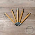 Set of wooden pens on a wooden surface with a brand logo in the corner.