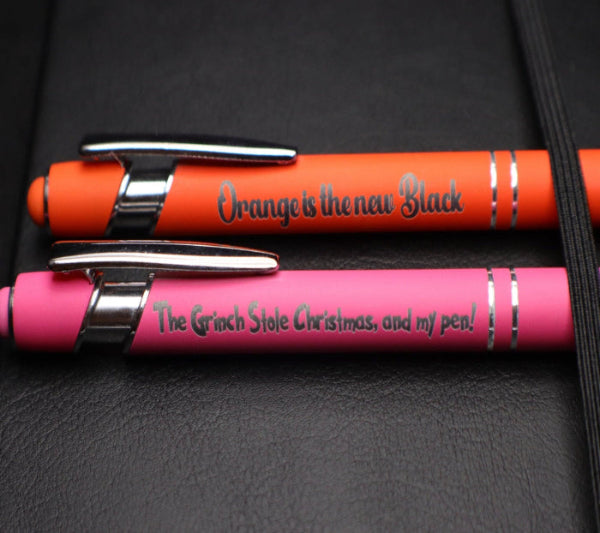 Two pens, one orange and one pink, with engraved text on a dark surface.